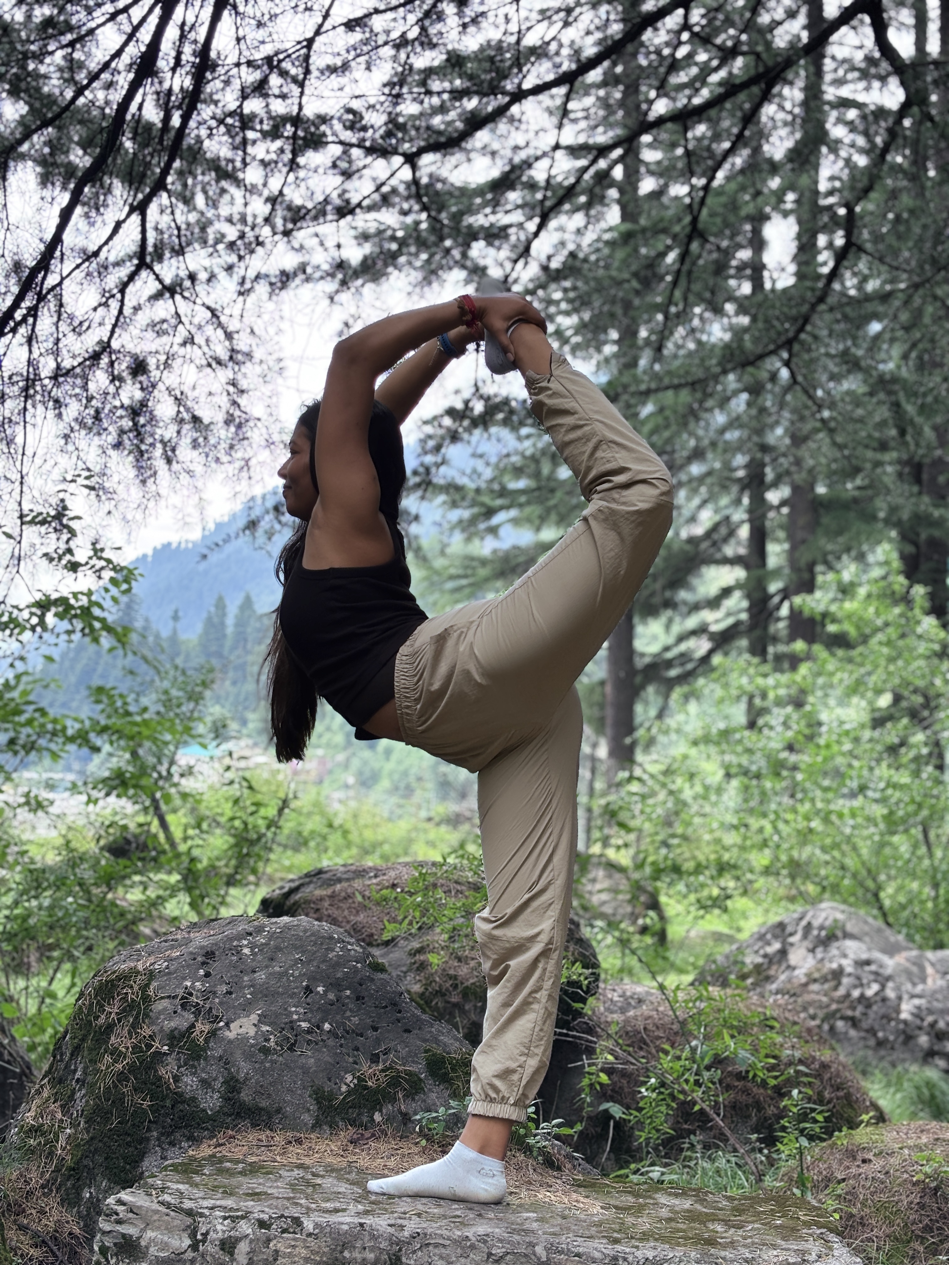 Purnima in Dancer Pose, a graceful standing backbend, on a rock amid mountain pines